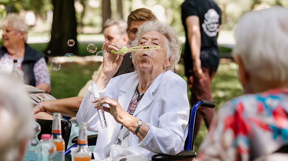ältere Frau draußen in Gruppe am Tisch bläst Seifenblasen
