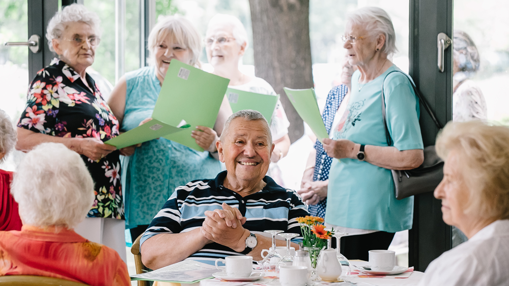 Senioren am Kaffetisch hinter ihnen stehend Seniorinnen mit Mappen