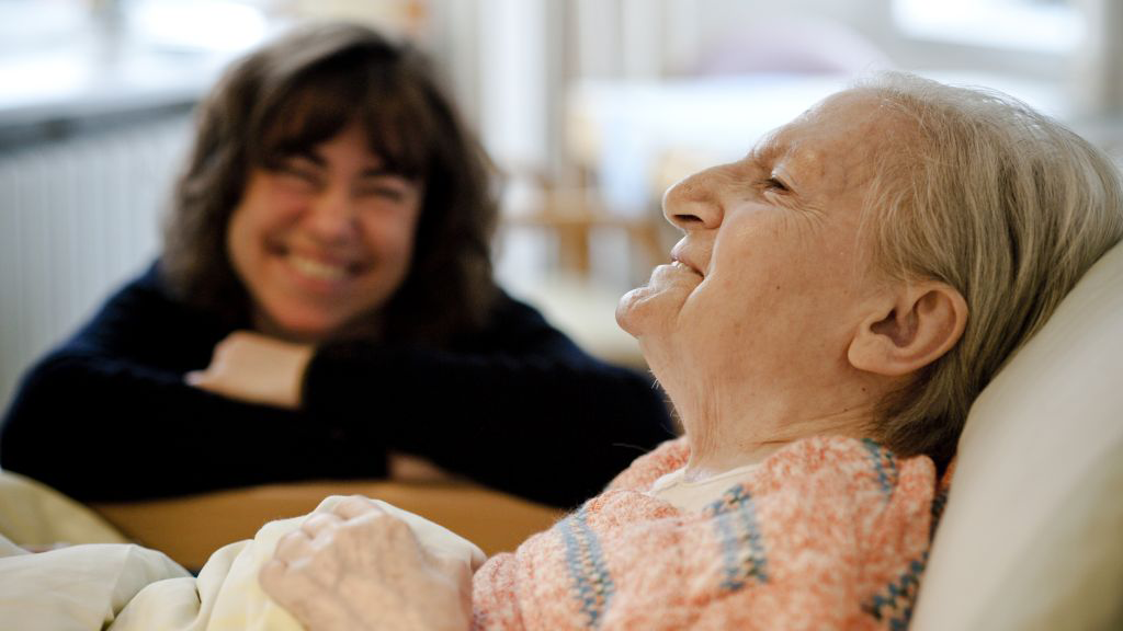 ältere Frau liegend lächelnd im Hintergund jüngere Frau lächelnd