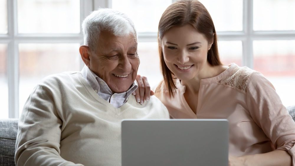 Ein älterer Herr und eine junge Frau schauen gemeinsam lachend auf einen Laptop.