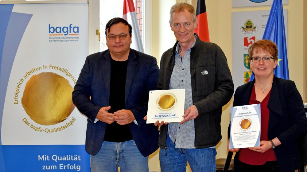 3 Personen stehen vor einem Banner, zwei von ihnen halten ein Zerfifikat in den Händen.